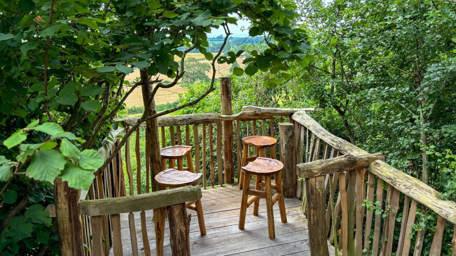 The rustic wooden treehouse deck in One Acre Wood is enveloped by lush green foliage. It features four natural wood stools, offering a serene view of the distant landscape and fields.