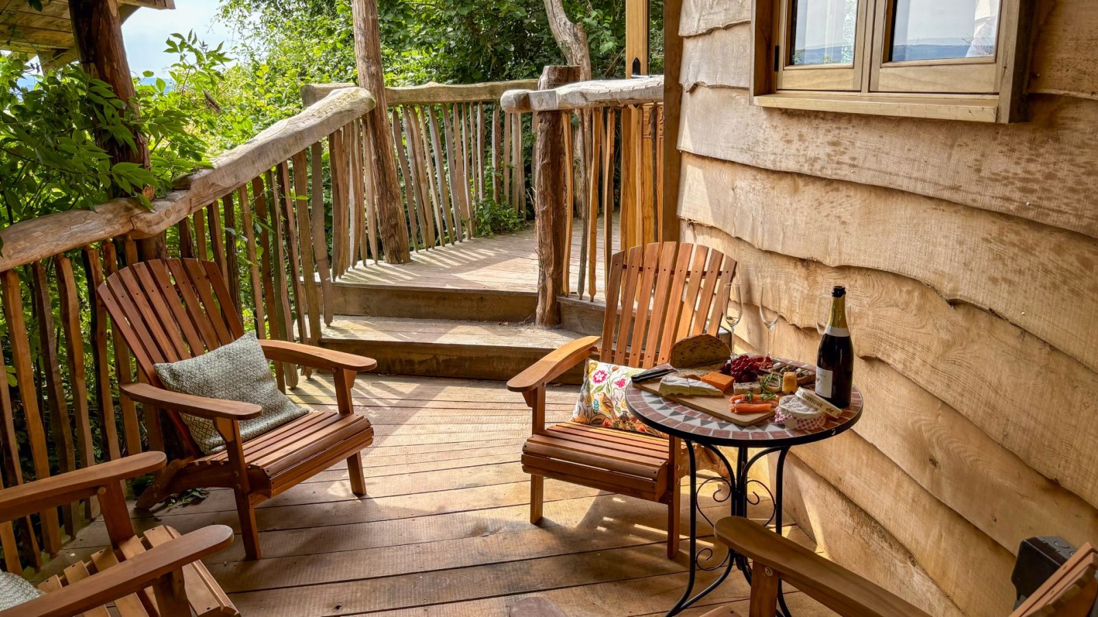 A cozy wooden deck set in the heart of One Acre Wood features two charming chairs with a small round table between them. The table holds a platter with snacks, a fresh loaf of bread, and a bottle of wine. Embraced by lush greenery and rustic wooden railings, it's an idyllic retreat.