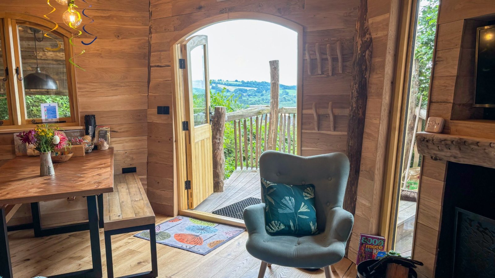 A cozy wooden interior reminiscent of One Acre Wood features an open door leading to a deck overlooking a scenic landscape. A cushioned chair with a leaf-patterned pillow sits near a small table brimming with flowers, while a colorful rug graces the entrance, bathed in natural light.