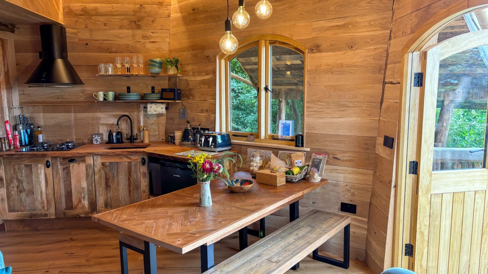 The rustic wooden kitchen interior at One Acre Wood features a wooden dining table with benches, a black stove, shelves stocked with dishes, and a bouquet of flowers. Hanging pendant lights illuminate the space, while large windows offer a peaceful view of the surrounding greenery.