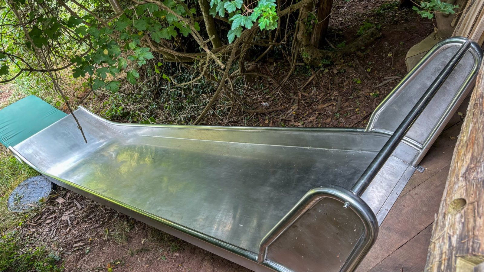 A shiny metal slide in One Acre Wood, surrounded by trees and bushes. The slide features green padding at the top with a dirt path winding nearby, creating a natural playground environment.