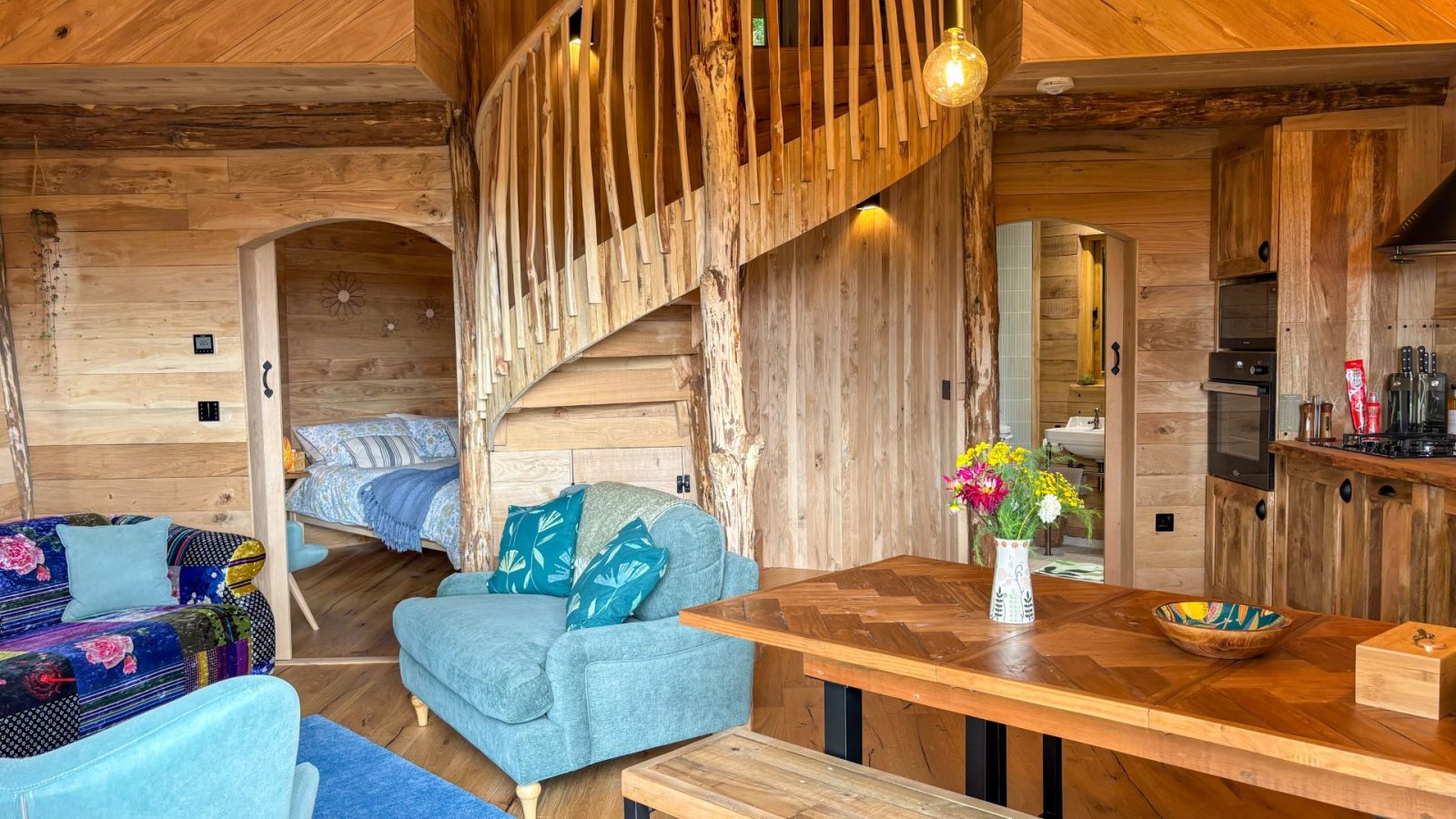 The cozy, rustic interior of One Acre Wood features wooden walls, a spiral staircase, and a small kitchen. Through an open door, a bedroom with a bed is visible. The living area boasts blue armchairs and a patterned sofa, while a table holds a vase with colorful flowers.