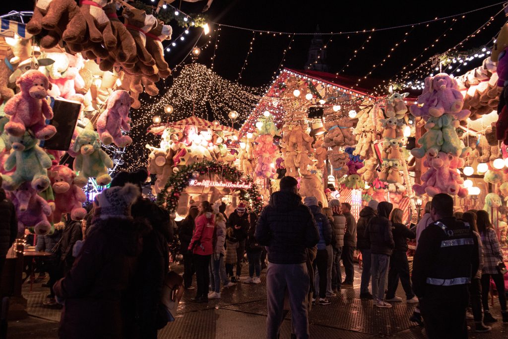 A festive night market, often cited among the Best Xmas Markets UK, sees people browsing and socializing. The market stalls are adorned with colorful, hanging plush toys, and twinkling string lights overhead create a warm and lively atmosphere.