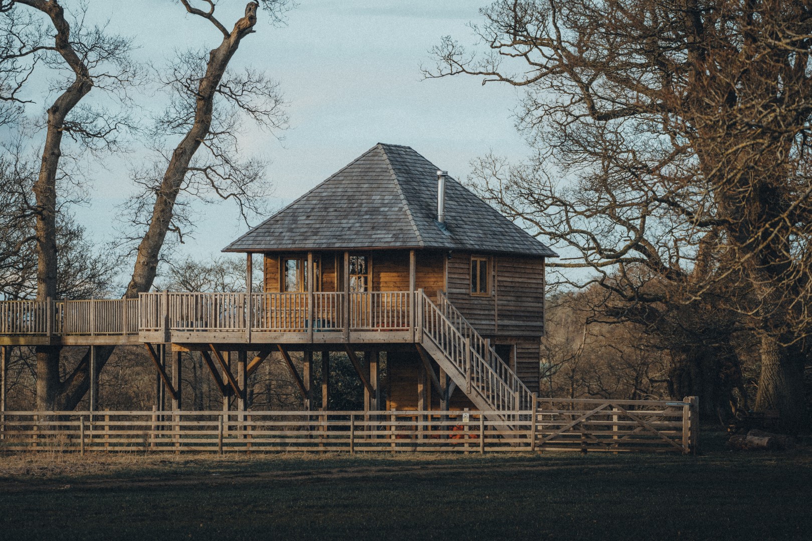 Treehouse with a wooden fence, elevated on stilts, nestled amidst leafless trees and a grassy area—perfect for those seeking luxury short breaks in the UK.