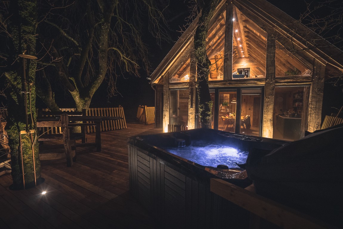 A cozy, rustic Prince Llywelyn Treehouse with large glass windows is illuminated warmly in the night. It features a wooden deck with a hot tub emitting a soft blue light. Surrounding the treehouse are large trees, enhancing the serene, nature-filled atmosphere.