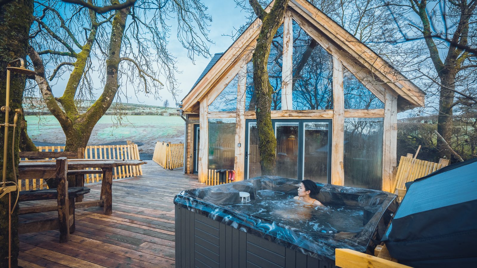 A cozy cabin with large windows, reminiscent of a unique treehouse stay, sits amid bare trees. A woman relaxes in a hot tub on a wooden deck, surrounded by this rustic escape. Nearby, a wooden table and lounge chairs offer a scenic view of the field in the background.