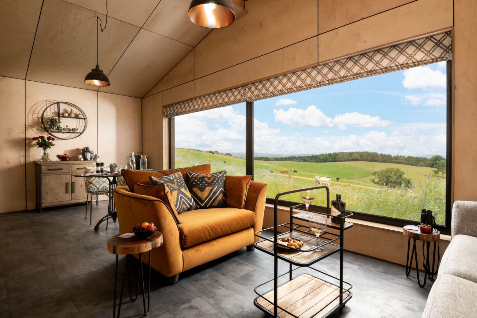 A modern living room with a large window offering a scenic view of rolling green hills and blue skies. Inside, there's a mustard yellow couch with patterned pillows, a side table with snacks and drinks, a hanging light fixture, and a stylish bar cart that makes this home feel like a picture-perfect page.