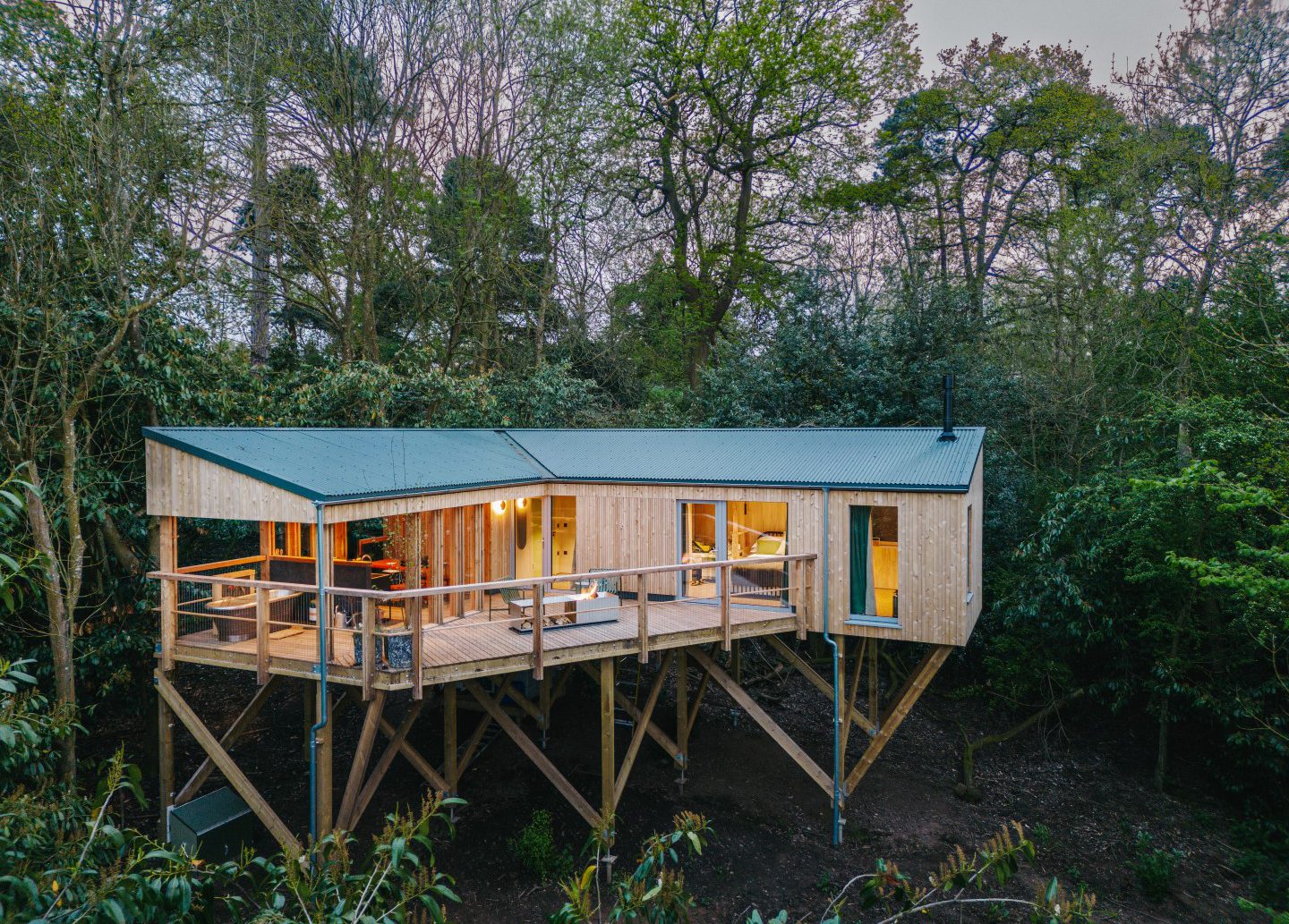 An elevated wooden treehouse nestled among tall trees, perfect for the home page of a nature lover's retreat. The structure features large windows, a wraparound deck with railings, and a green pitched roof. Warm light emanates from inside, creating a cozy and inviting atmosphere amidst the lush greenery.