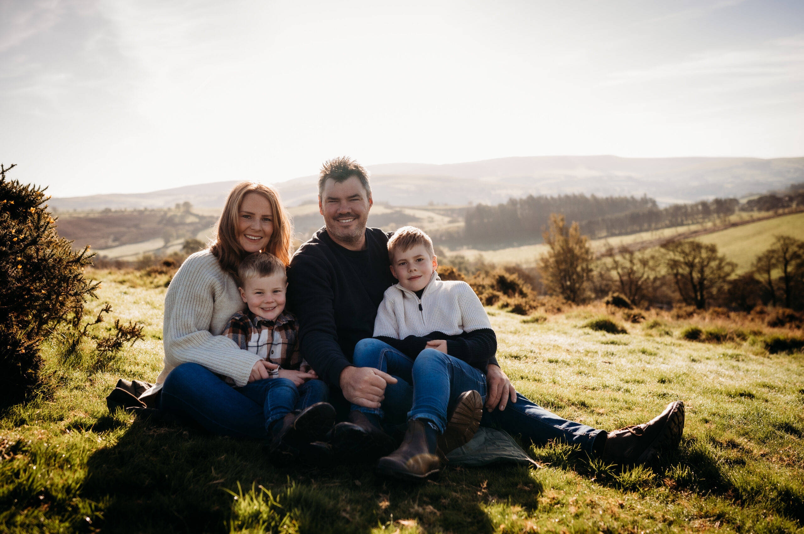 A family of four sits on a grassy field with rolling hills and trees in the background. The parents are seated in the middle, smiling, with their two young children sitting on either side. Dressed in casual, warm clothing and basking under the bright sun, they look like the perfect feature for a travel blog by ToWander.