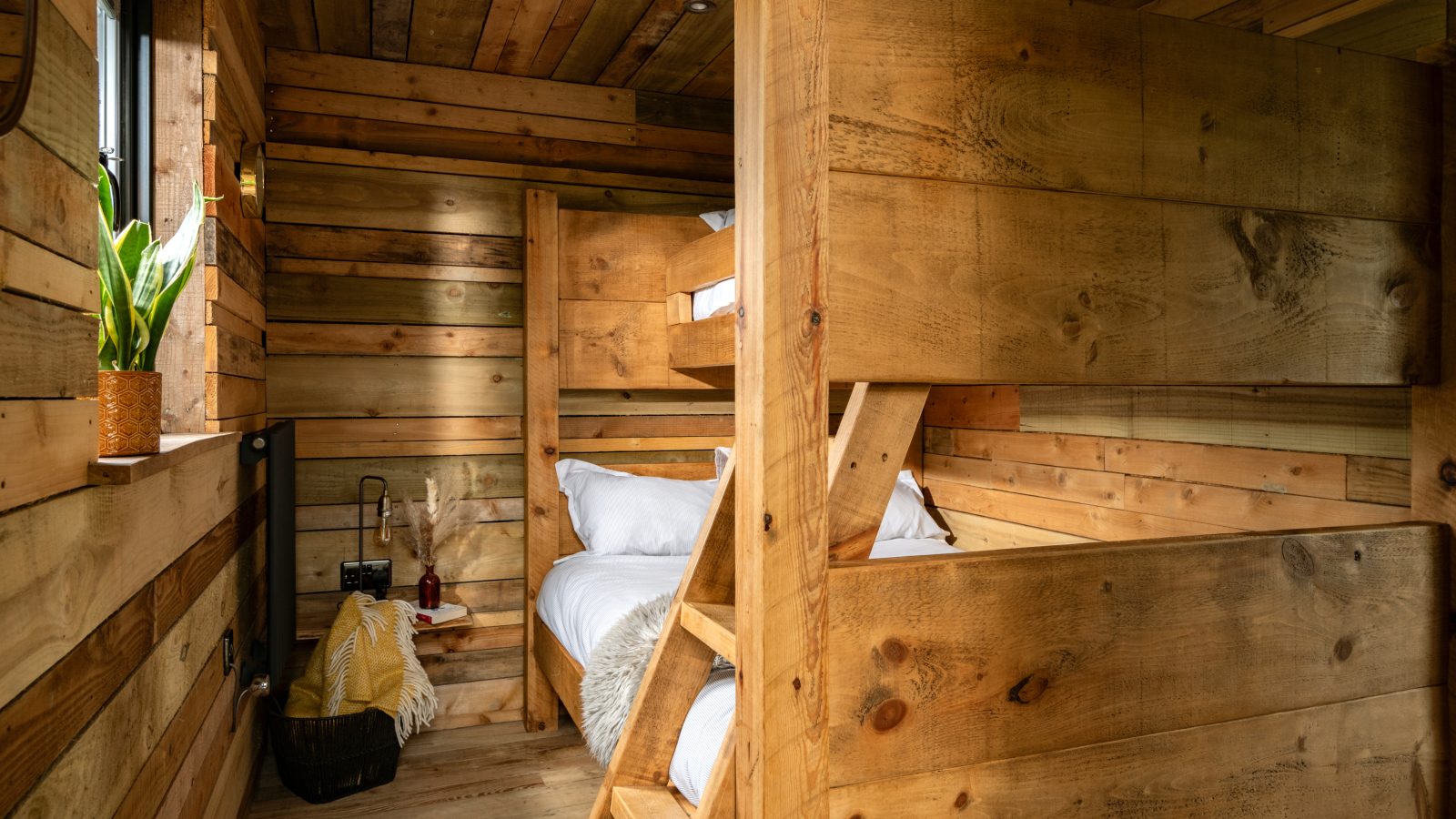 A cozy cabin room reminiscent of a treehouse, with wooden interior walls and a bunk bed. A small window lets in natural light, highlighting a potted plant on the sill. A basket with a blanket and a decorative vase adds warmth and charm to the space.