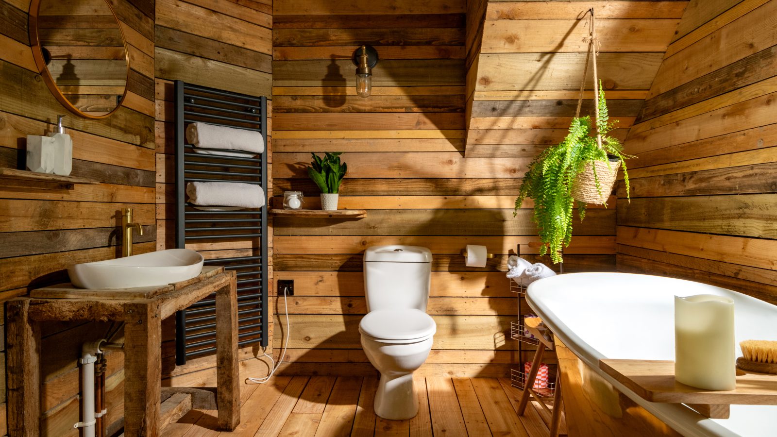 A rustic bathroom in the Prince Llywelyn Treehouse features wooden walls and flooring, a bathtub, a white toilet, and a wooden sink counter. A round mirror hangs above the sink, while towels warm on a heated rack. A hanging plant adds greenery as natural light illuminates this Welsh accommodation.