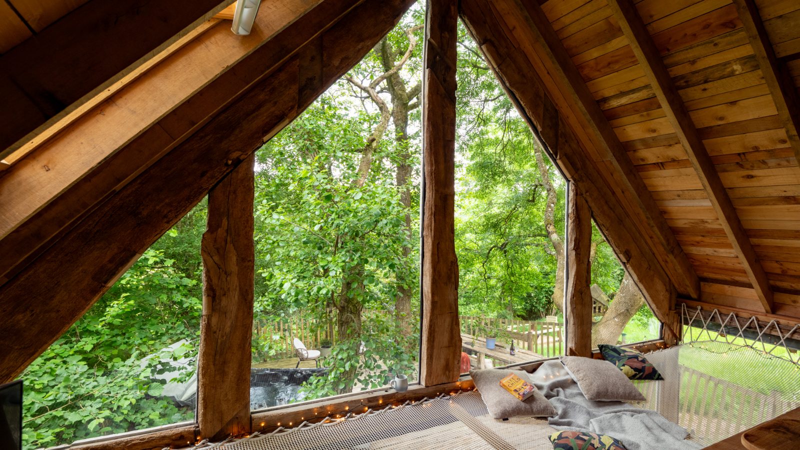 Cozy attic with large triangular windows offering a view of lush green trees, reminiscent of a luxury treehouse. The space features a wooden ceiling and hammock-style floor, decorated with pillows and a blanket for a relaxing, nature-inspired retreat. Perfect for treehouse accommodation enthusiasts.