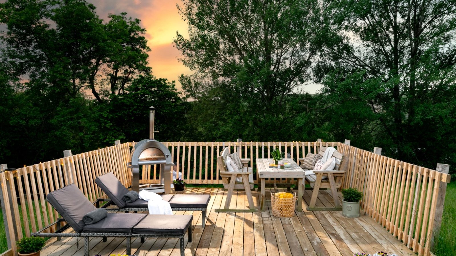 A wooden deck with cushioned lounge chairs and a dining table set is surrounded by a fenced railing, reminiscent of the cozy charm of a Prince Llywelyn treehouse. A wood-fired oven stands in the corner. Green trees and a sunset sky create a serene backdrop, while potted plants and flowers decorate the space.