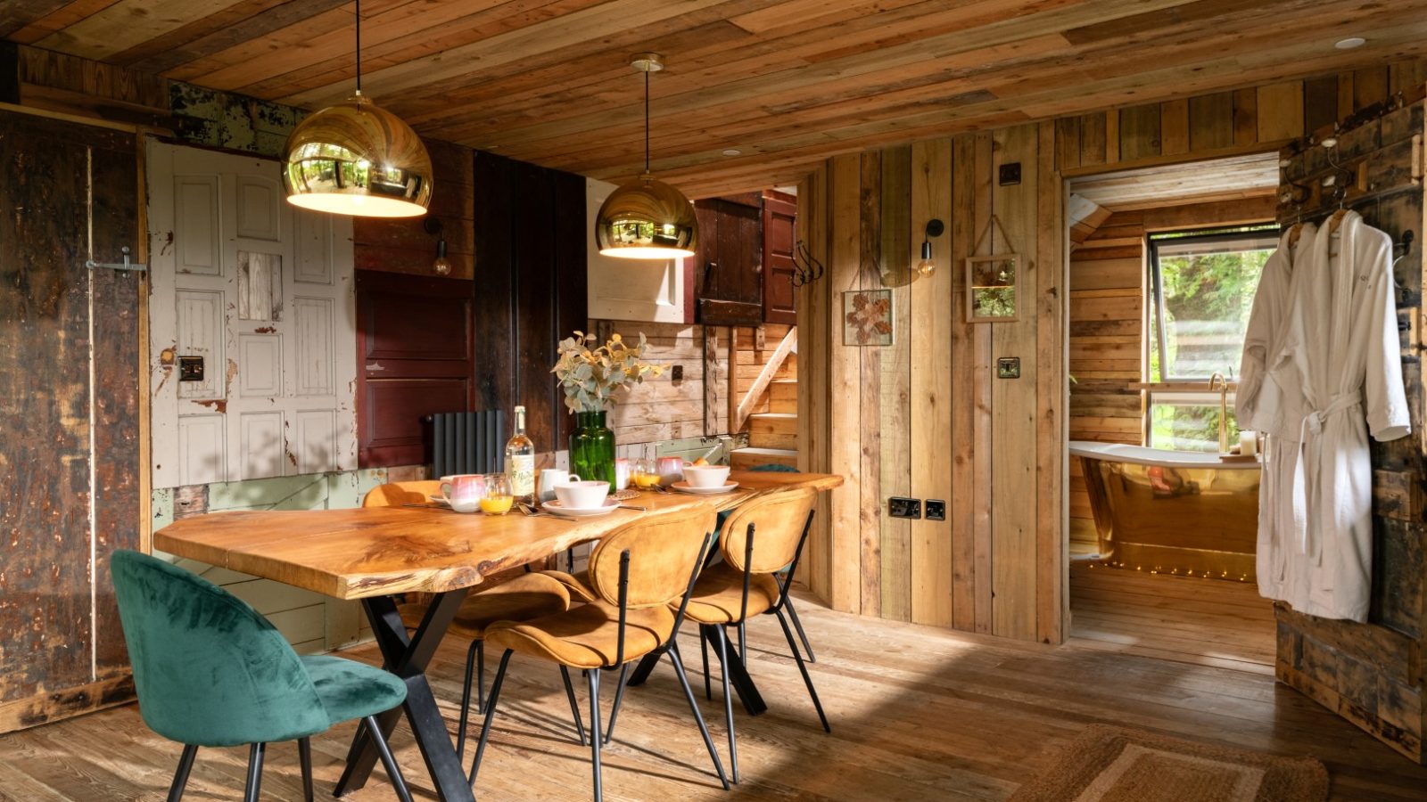 Cozy dining area with a wooden table and chairs, two golden pendant lights, and rustic wood-panel walls reminiscent of a treehouse. A green chair adds contrast. A doorway leads to a room with a freestanding bathtub fit for Prince Llywelyn. Decor includes flowers and a hanging robe.