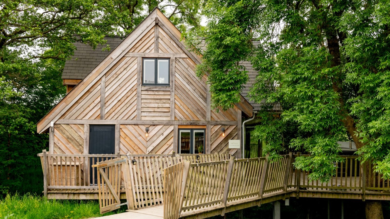 A rustic wooden cabin with a slanted roof is surrounded by lush green trees, reminiscent of a cozy treehouse. It features a wooden ramp leading to the entrance and an upper-story window. The cabin, evoking the spirit of Prince Llywelyn, is nestled in a peaceful, natural setting.