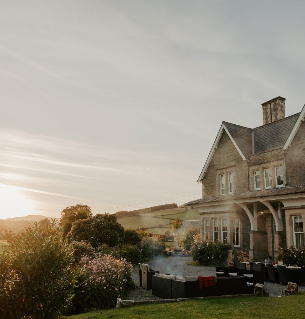 A large, vintage-style house with a stone exterior and pitched roof is surrounded by lush greenery. Outdoor seating is arranged on the patio, perfect for potential buyers wanting to list with us. The sun sets in the background, casting a warm glow over the scene as smoke rises from a barbecue.