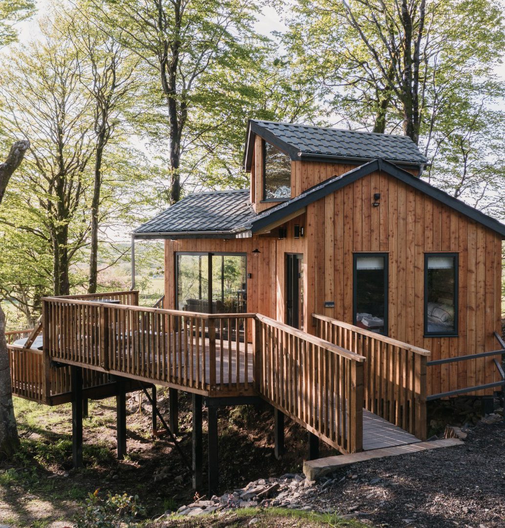 A small wooden cabin with large windows and a wraparound deck is nestled among tall, leafy trees. The modern, rustic structure is elevated on stilts with a pathway leading to the entrance, blending seamlessly into its lush forest surroundings. List with us to make this serene retreat your own!