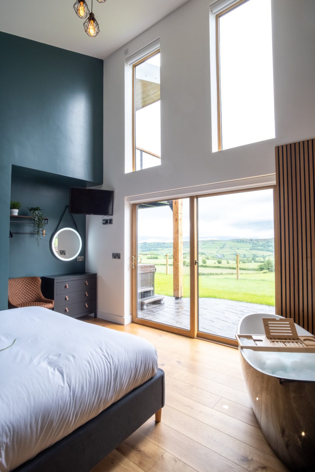 A modern bedroom with floor-to-ceiling windows showcasing a scenic view of rolling hills. The room features a cozy bed, a vanity with a round mirror, a chair, and a freestanding bathtub near the sliding glass doors. The interior decor is contemporary with wooden elements, reminiscent of luxurious home page designs.