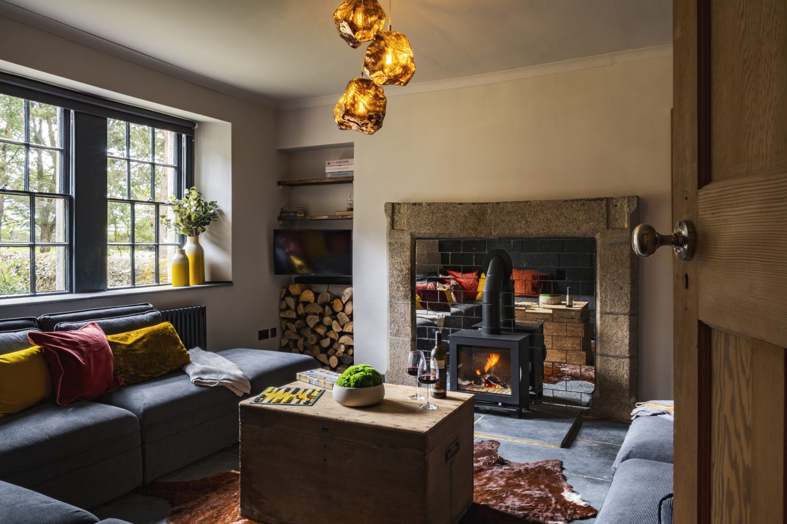 A cozy living room featuring a wood-burning stove with a stone backdrop, surrounded by a sectional gray couch adorned with colorful pillows. A wooden coffee table with decor sits on a cowhide rug. The room, perfect for those who love to wander, is illuminated by unique amber pendant lights and large windows.