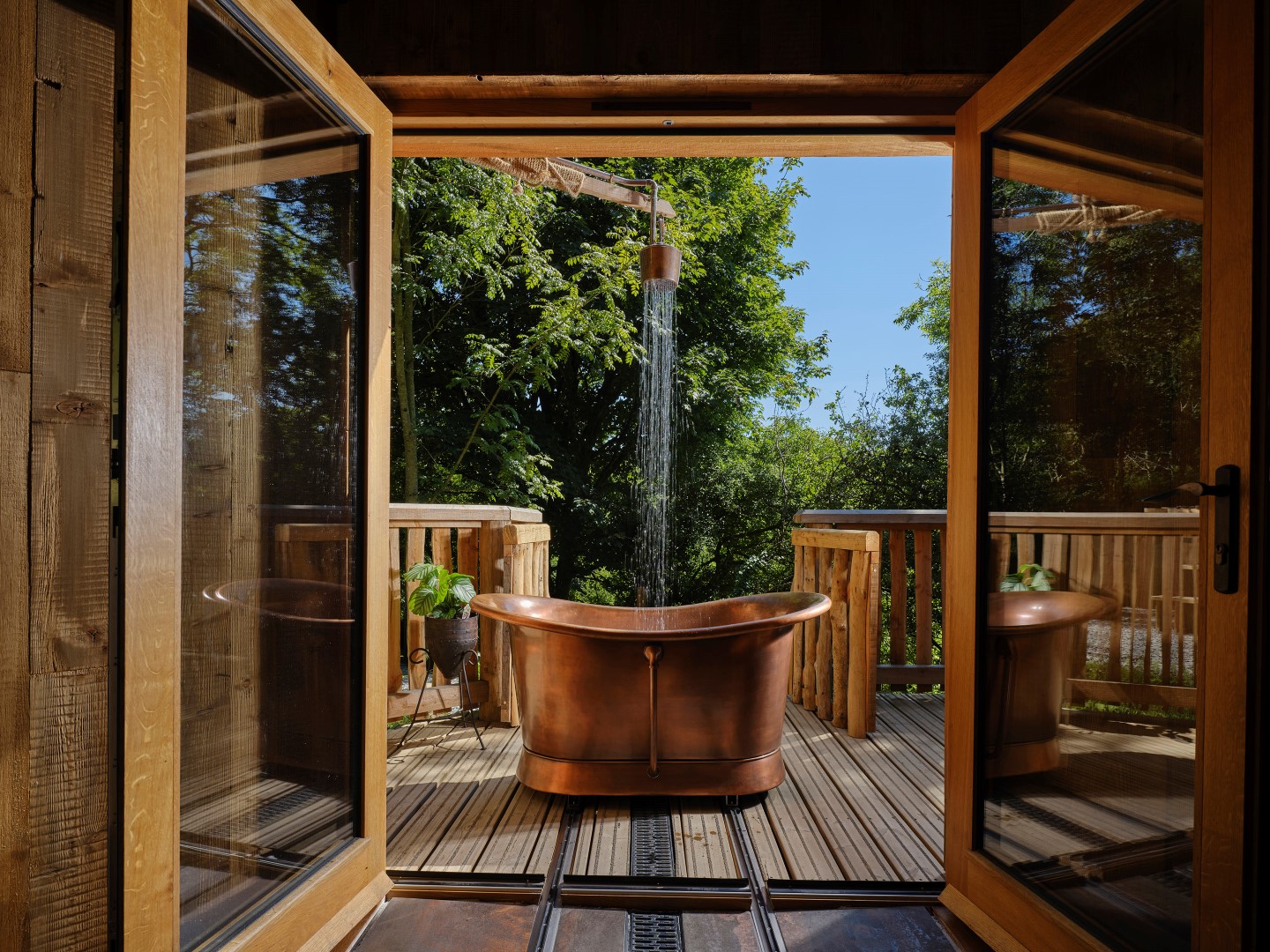 A rustic outdoor bathroom with a freestanding copper bathtub on a wooden deck, surrounded by lush greenery. A rain shower head is positioned above the tub. Glass doors open up to this serene home sanctuary, creating an airy atmosphere amid nature that feels like a page from a dream.