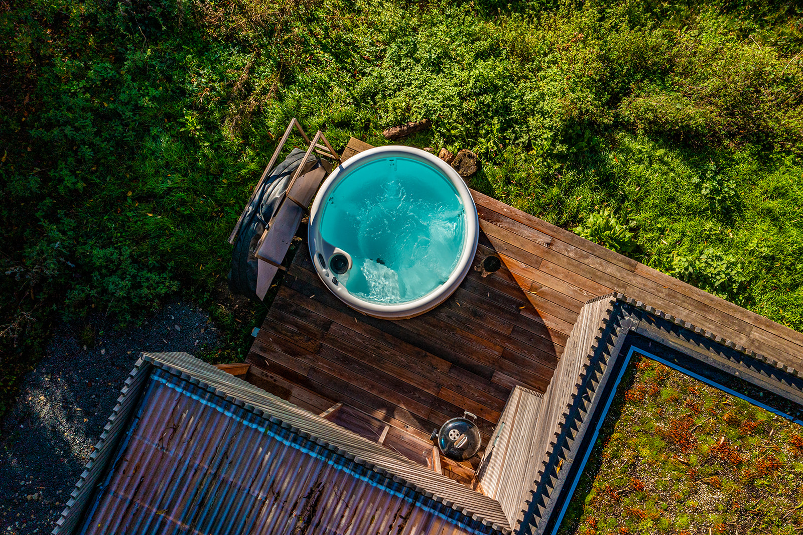 Aerial view of a hot tub filled with water on a wooden deck beside a modern building. The surrounding area is covered with lush green vegetation, creating a natural and serene environment. A black stool is placed next to the hot tub. Experience this slice of paradise when you list with us.