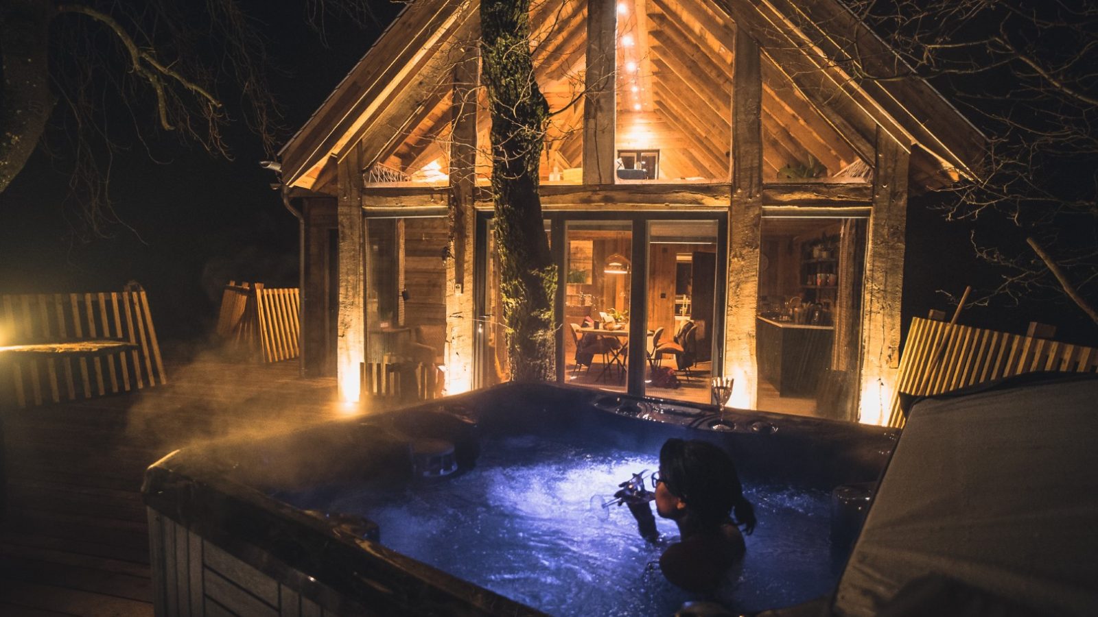 A person relaxes in a steaming hot tub on a wooden deck at night. Warm, inviting lights illuminate the Prince Llywelyn Treehouse, with large glass windows in the background, creating a cozy atmosphere. This unique stay promises an unforgettable treehouse accommodation experience.