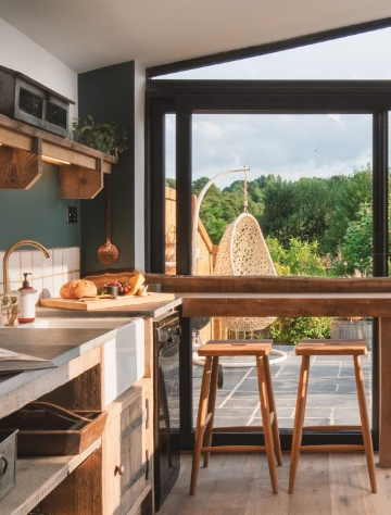 A cozy kitchen features rustic wooden countertops and shelves. Two wooden stools sit by a large window that opens to a sunny patio with a hanging wicker chair. Outside, lush greenery and a serene landscape enhance the tranquil, inviting atmosphere of this home.