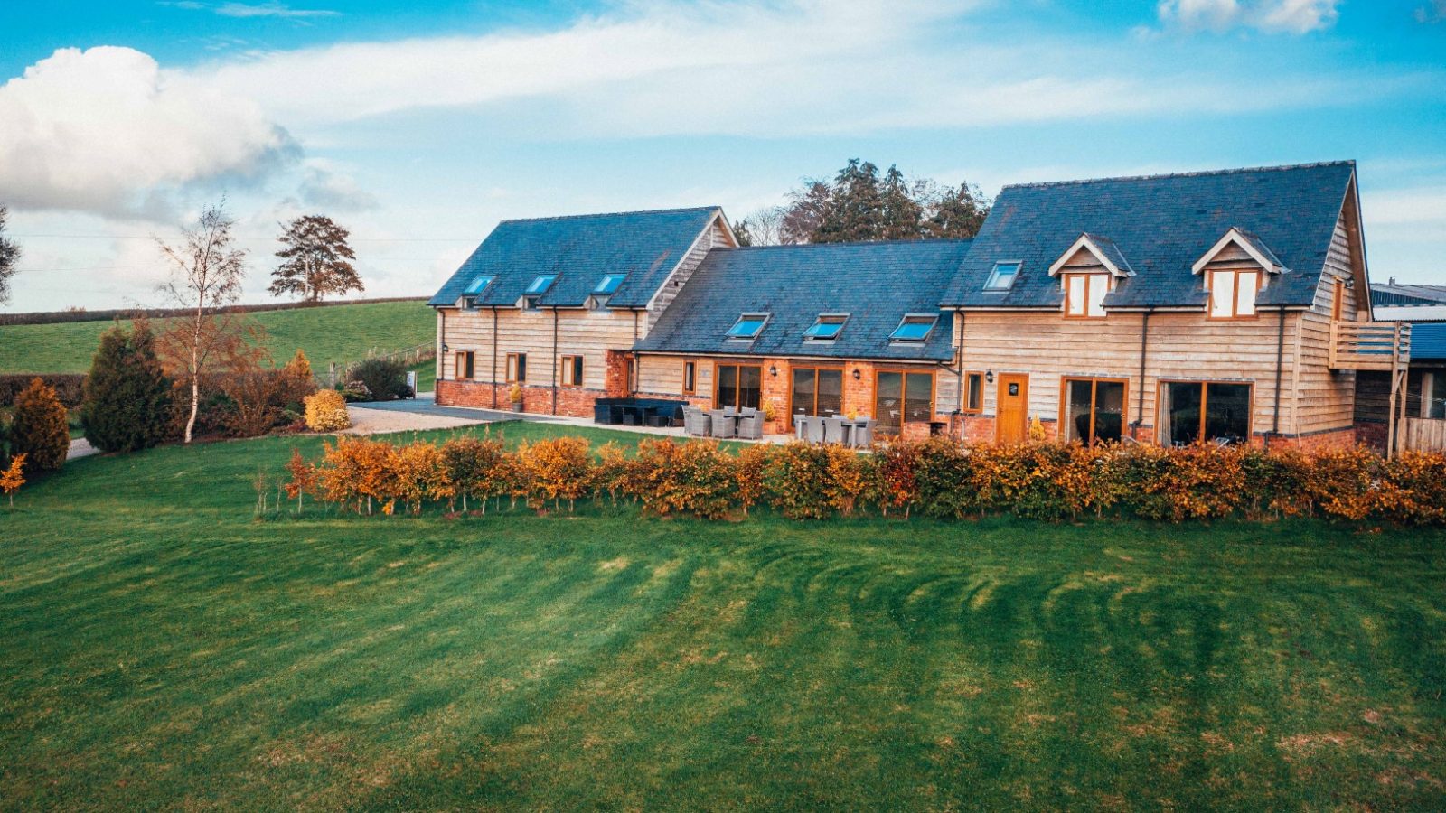 A unique, large house with wooden siding and a slate roof, set against a rural Welsh landscape. The manicured lawn and hedges complement the clear sky with its few clouds, while trees stand proudly in the background—a perfect spot for memorable Welsh stays.