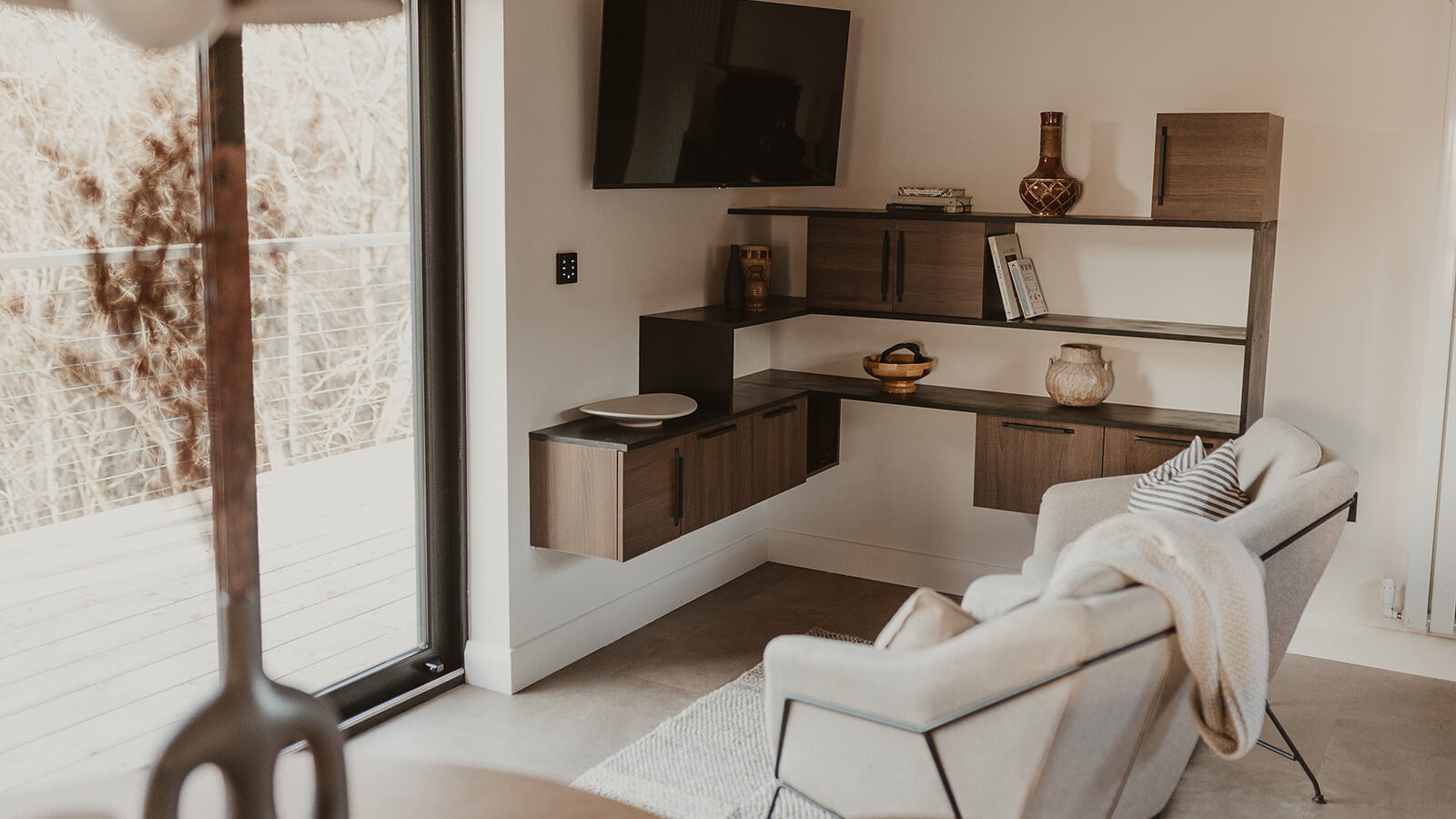 A modern living room with wall-mounted shelves, a flat-screen TV, and cozy seating near a large window offers a lodge-like warmth, perfect for enjoying serene views while nestled in comfort.