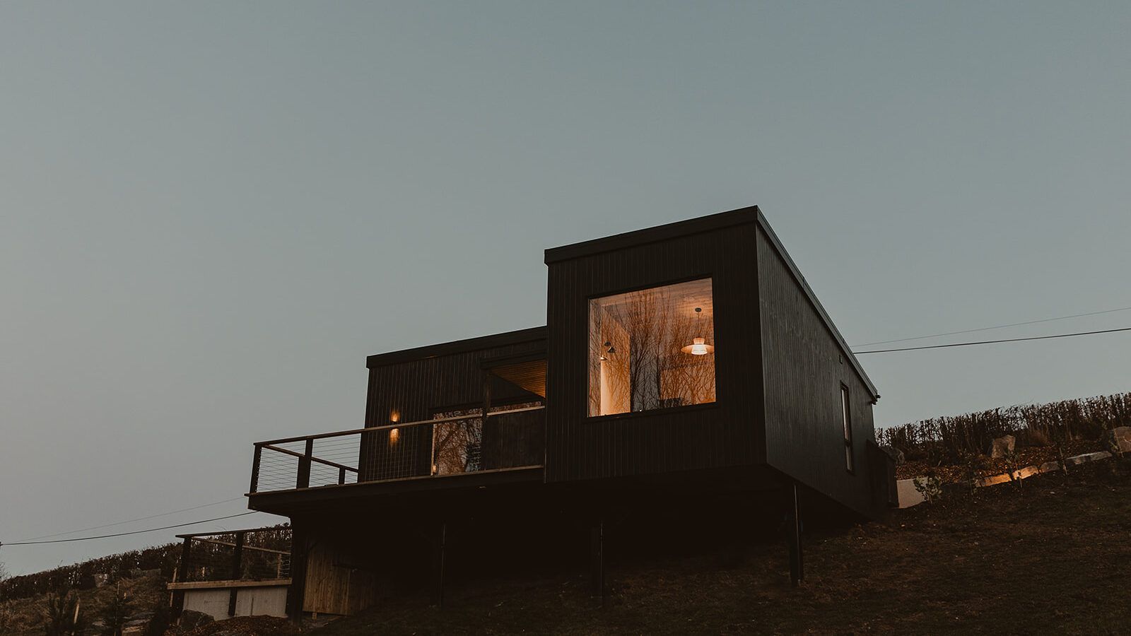 Modern black lodge with large windows and a deck, nestled on a hillside at dusk.