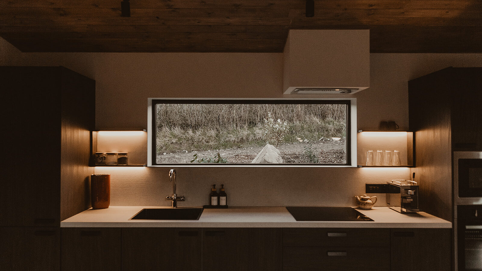 The minimalist kitchen at Berry Bush Lodge features dark wood cabinets, a white countertop, and a sink. A large window offers an expansive view of the outdoors, bringing a touch of nature into the serene lodge setting.