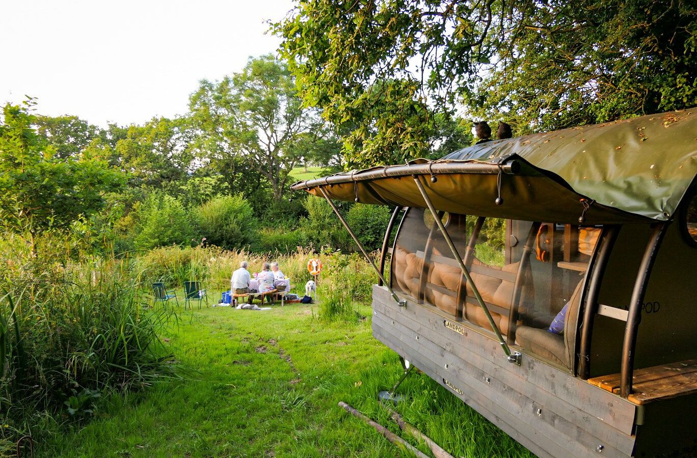 A group of people is seated at a picnic table near the wooded expanse of Coppet Hill, with a rustic wooden cabin on wheels nearby.