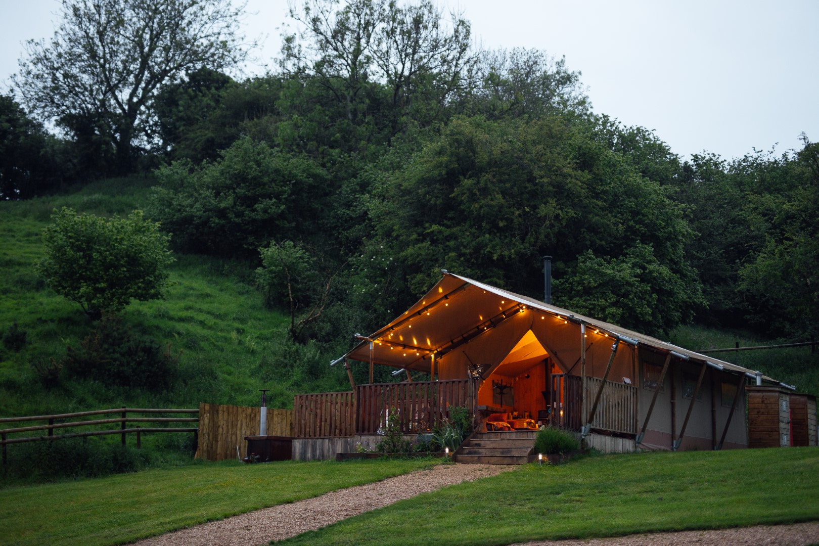 A warmly lit wooden cabin with a porch is nestled in lush greenery, surrounded by trees and a grassy hill, offering an idyllic retreat reminiscent of luxury safari tents.