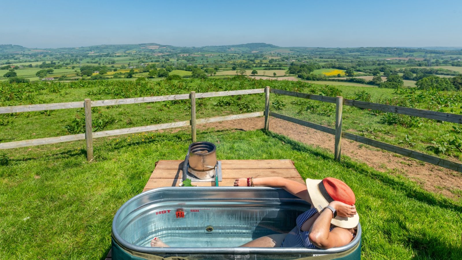 Person in a hat relaxing in an outdoor tub with a serene Coppet Hill countryside view.