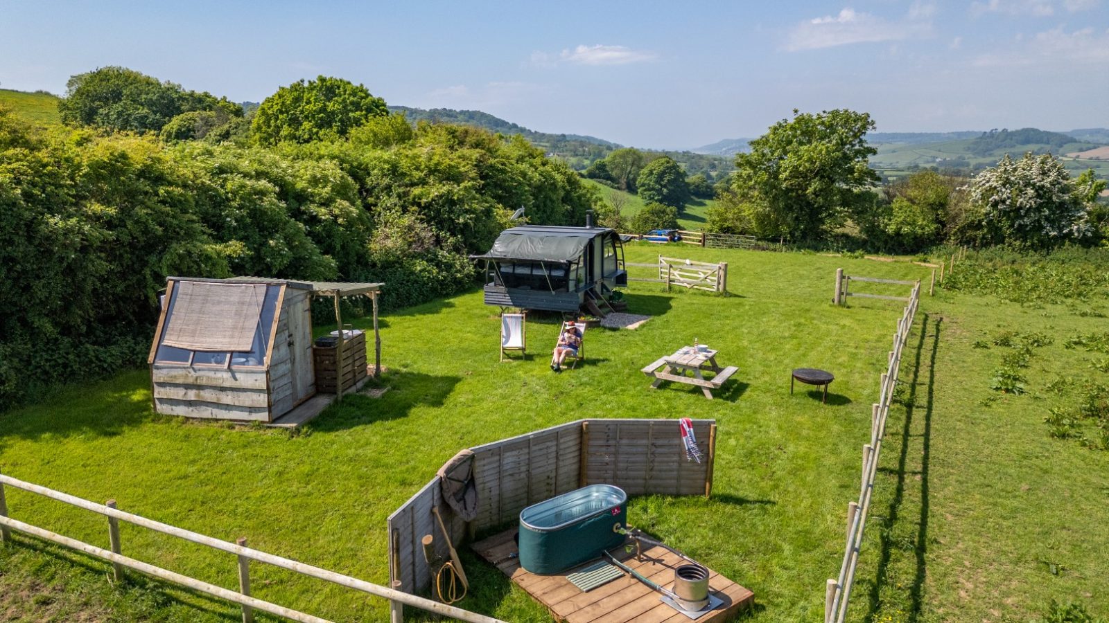 Aerial view of a fenced green lawn with a trailer, wooden structure, picnic table, and bathtub near Coppet Hill, surrounded by hills.