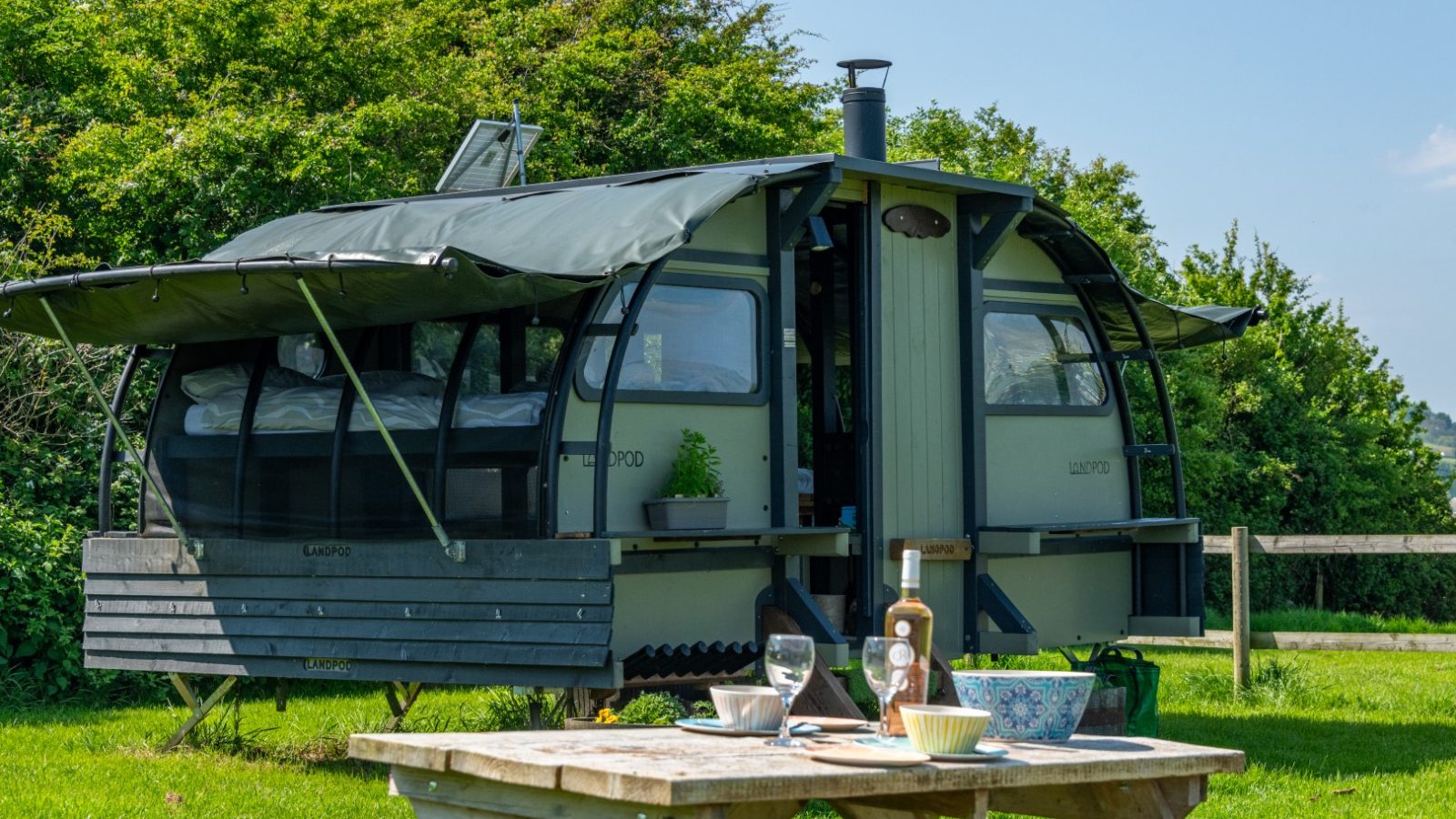 A tiny cabin with open windows and a canopy sits on Coppet Hill, with a picnic table under a clear sky on the grassy land.