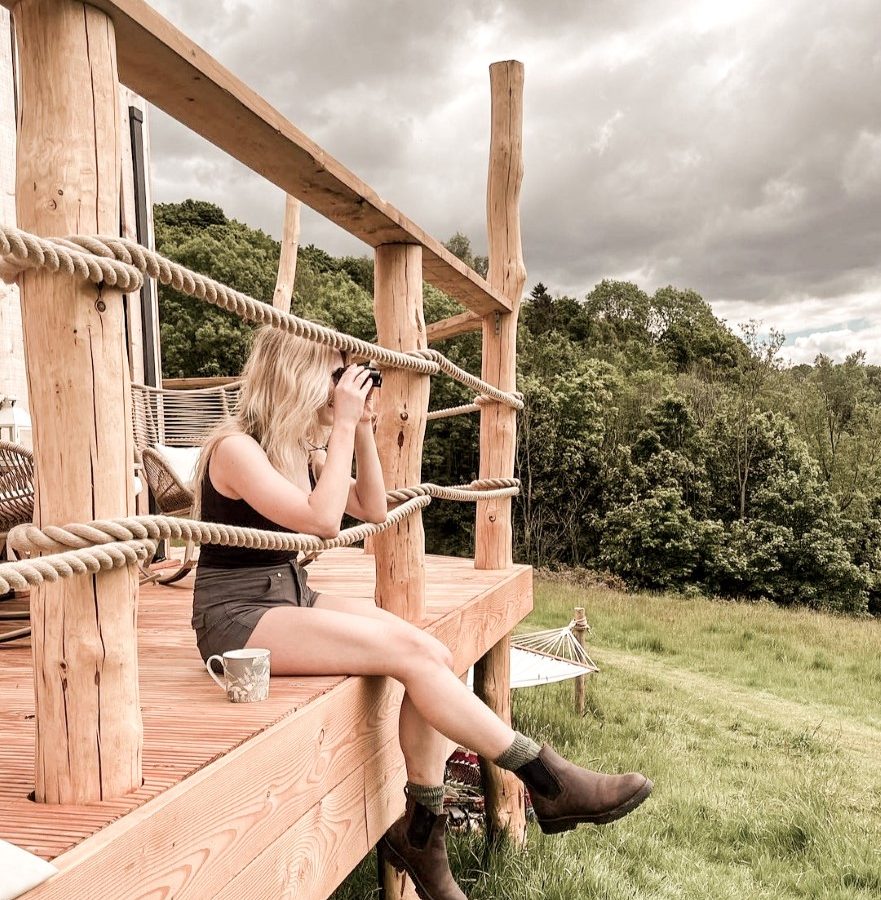 A woman sits on a wooden porch, using binoculars to admire the lush green landscape of the Peak District. The cloudy sky looms overhead. A mug is placed beside her, and a hammock hints at her quiet hideout in the background.