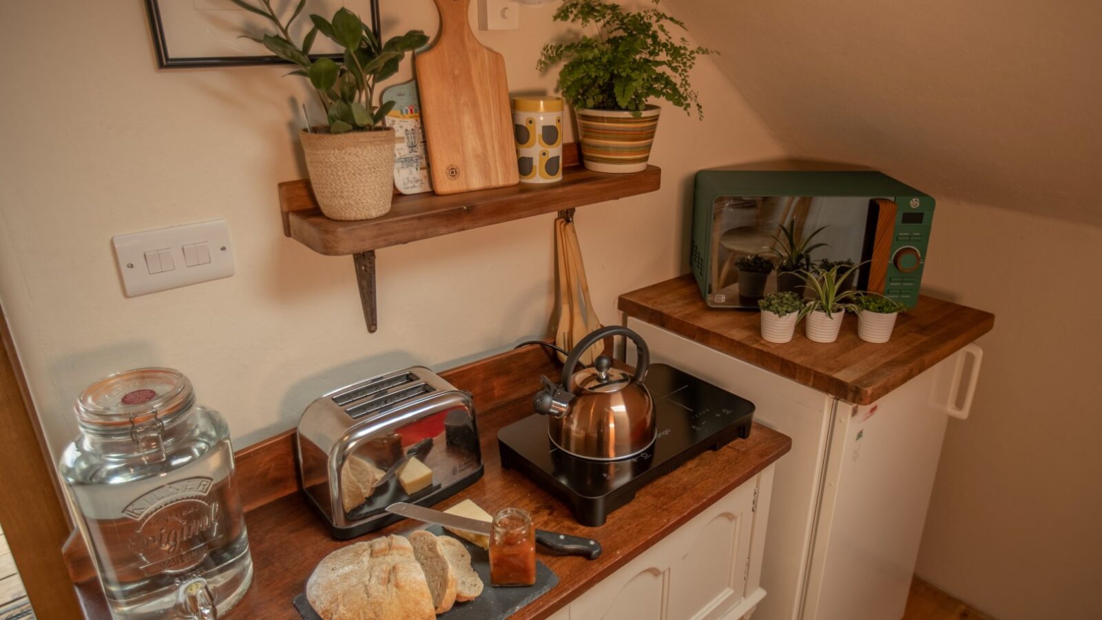 Cozy kitchen corner in your Snowdonia hideaway, complete with a toaster, sliced bread, a copper kettle, a green microwave, potted plants, and a small refrigerator.