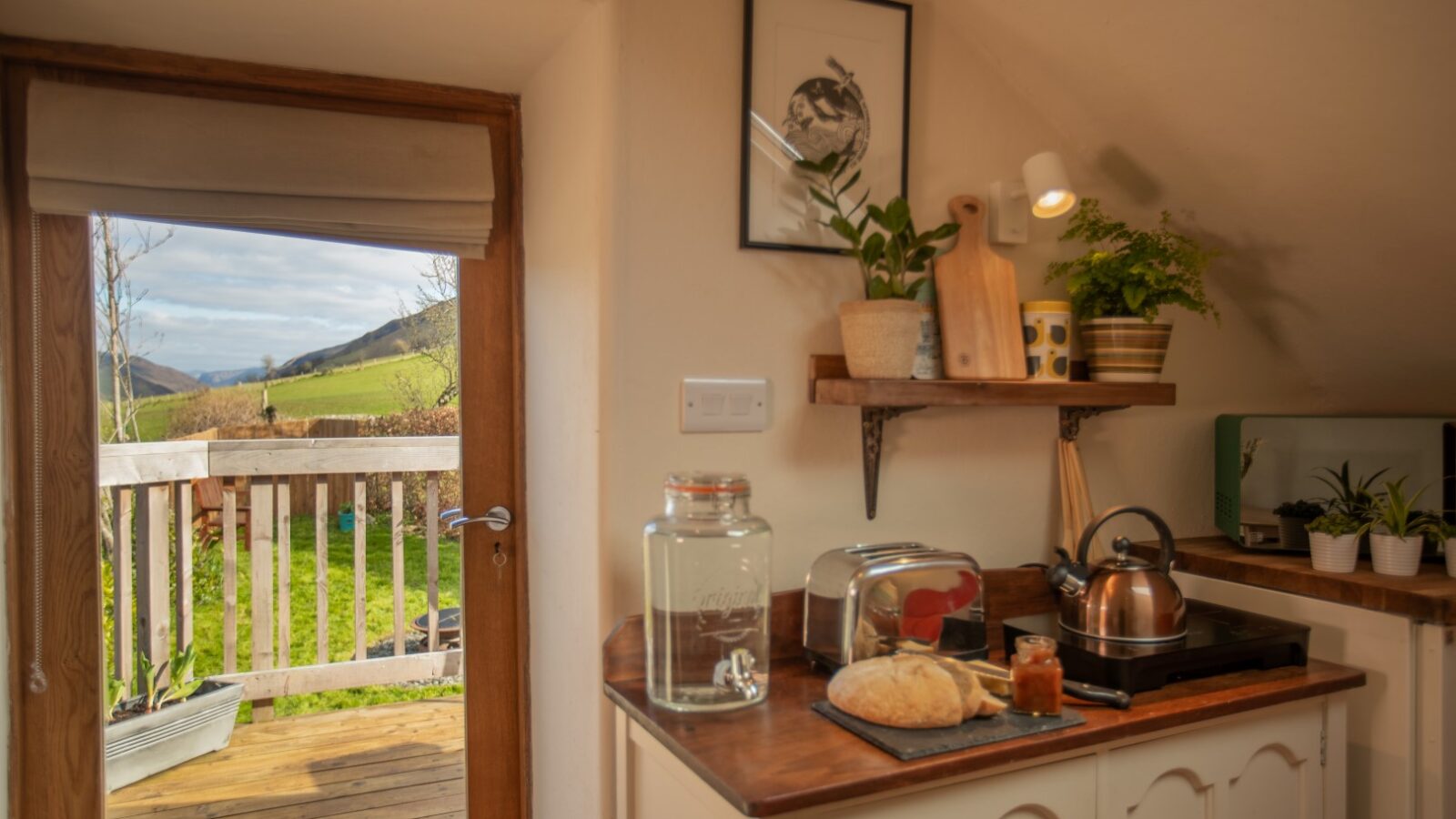 Cozy kitchen with bread, toaster, and kettle on the counter. Plants and cutting boards adorn the shelves. The doorway opens to a scenic Snowdonia hideaway, offering a tranquil retreat from everyday life.