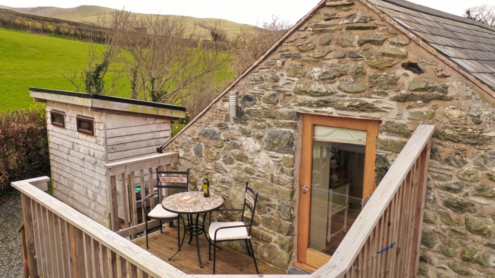 A stone cottage with a wooden deck featuring a table, two chairs, and a wine bottle is nestled in this Snowdonia hideaway, set against a rural backdrop.