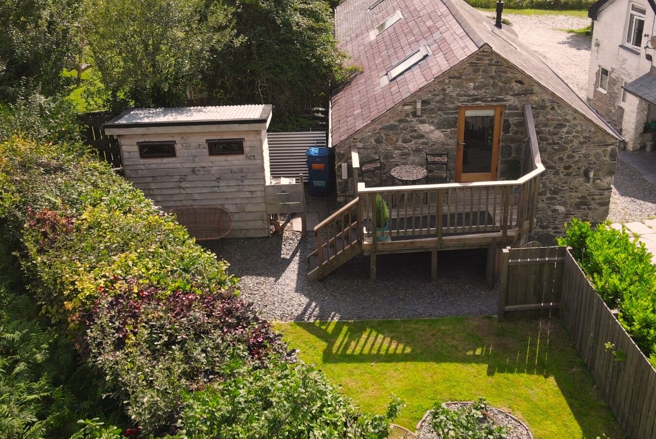 Aerial view of a charming stone cottage hideaway with a small wooden shed and garden, nestled in the lush greenery of Snowdonia on a sunny day.