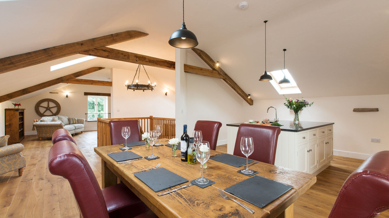 A spacious open-plan dining room in the holiday cottage features a wooden table, red chairs, and a modern kitchen under a vaulted ceiling with beams.