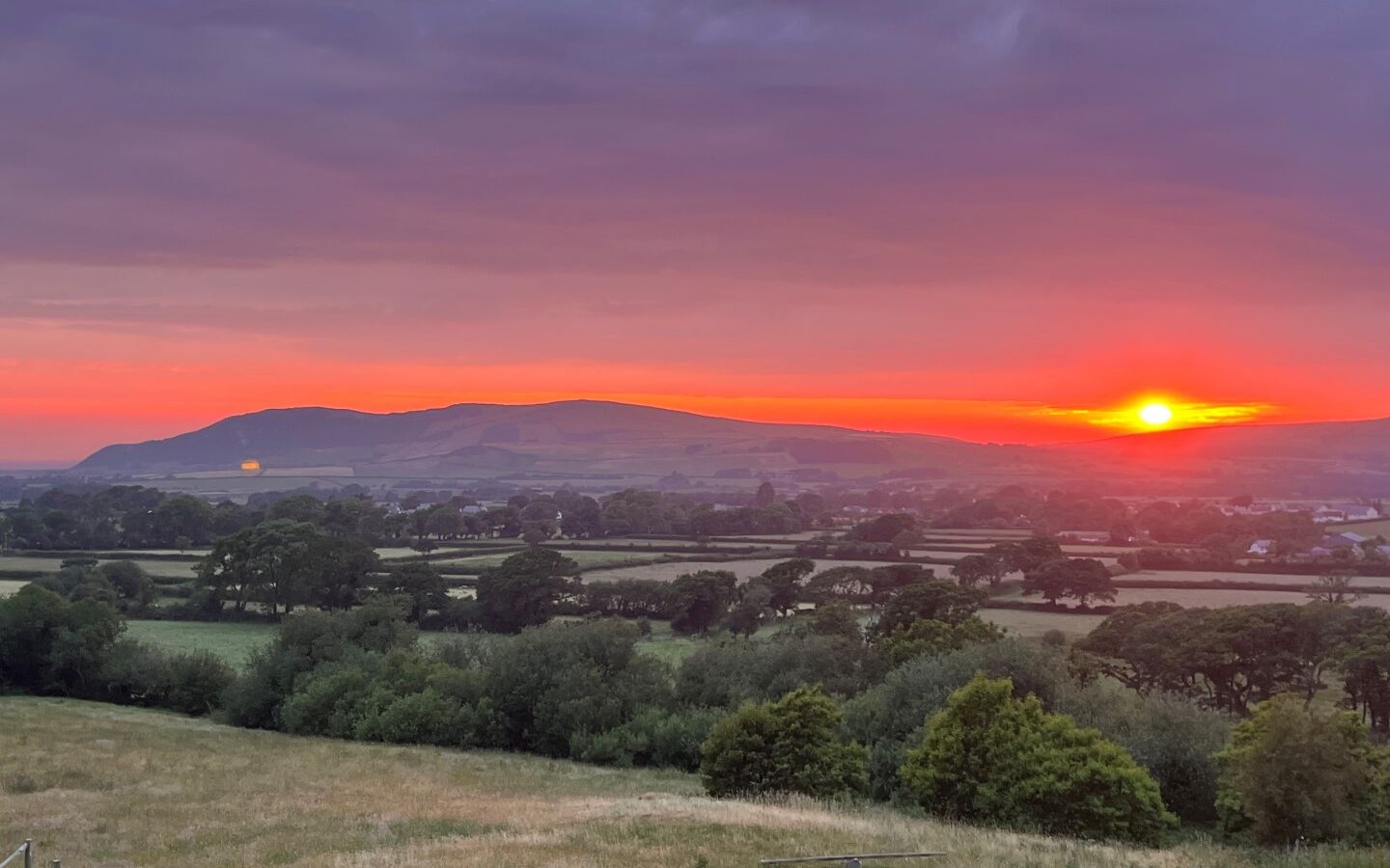 Sunset over a rural Snowdonia landscape with distant hills, fields, and trees under a colorful sky.