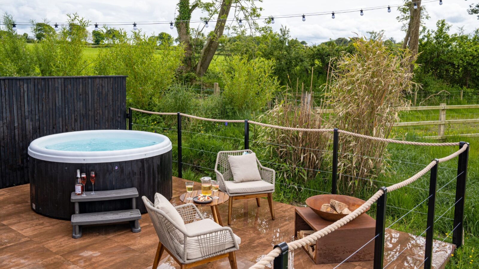 A charming outdoor patio at the Rusty Boathouse features a round hot tub, two wicker chairs, a table with drinks, and a cozy fire pit. String lights twinkle above as you enjoy this waterfront dining spot in a lush garden setting.