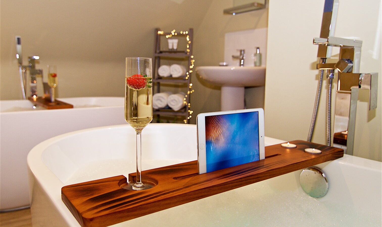 The Georges Getaway wooden bath tray gracefully holds a glass of champagne, a tablet, and flickering candles over a bathtub in a cozy bathroom setting.