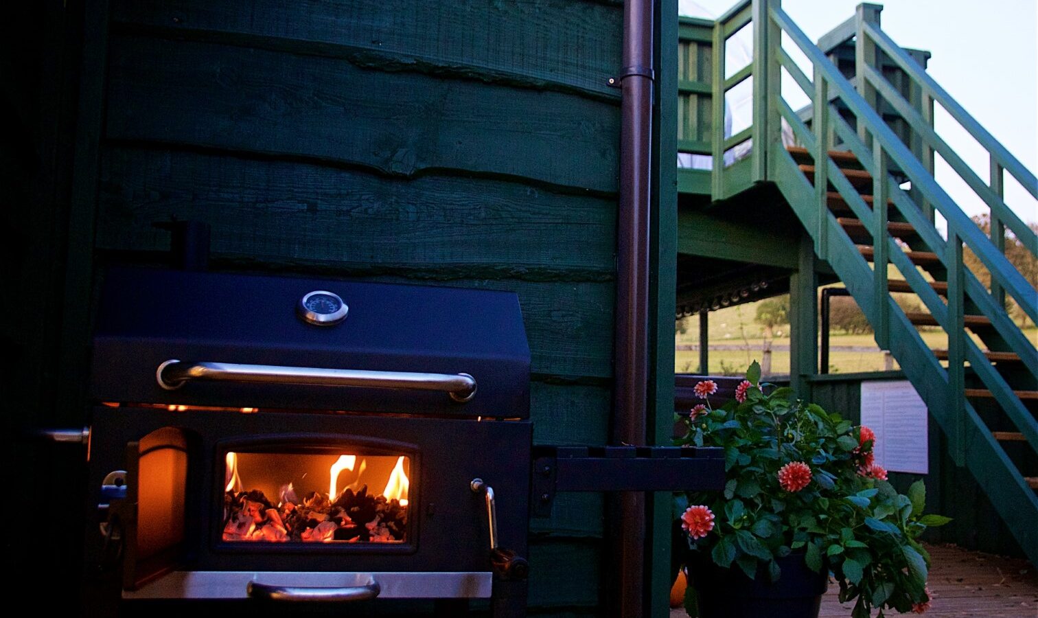 Georges Getaway features an outdoor wood-fired oven with visible flames next to a green wall and staircase, complemented by a potted plant bursting with red flowers.