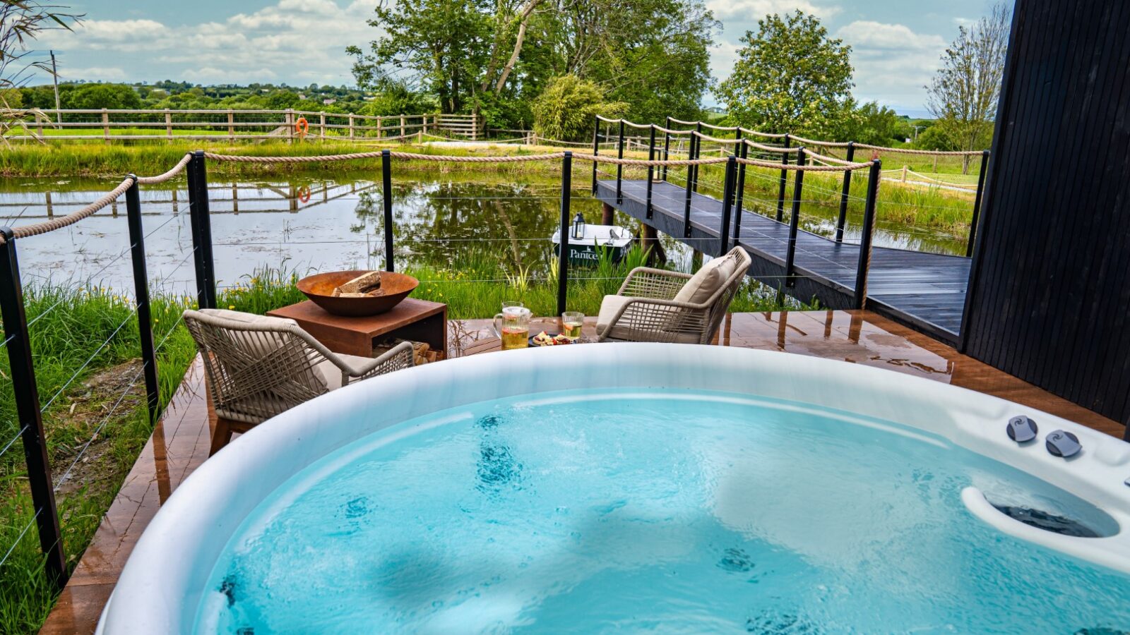 Hot tub overlooking a serene pond with wicker chairs, a wooden bridge near a rustic boathouse, and lush greenery under a partly cloudy sky.