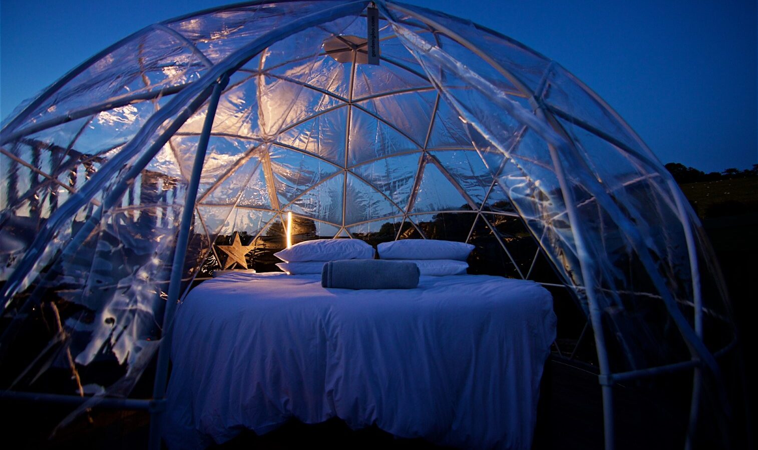 Georges Getaway: A clear dome structure with a cozy bed and pillows inside, illuminating at dusk against a dark blue sky.