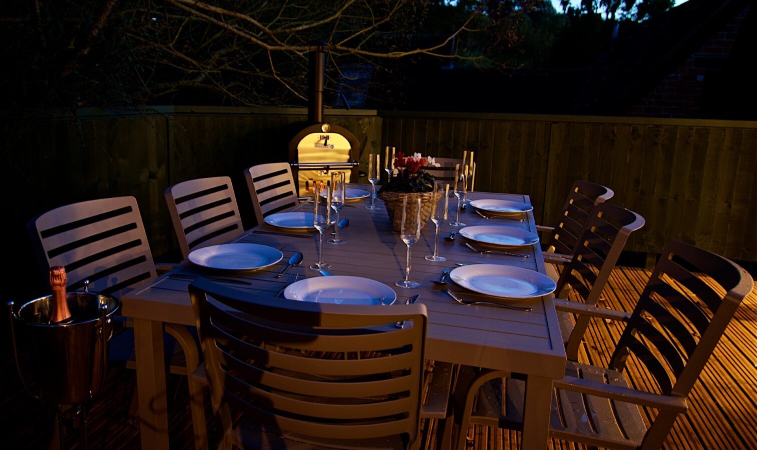 At Georges Getaway, enjoy an outdoor dining table set with elegant plates, glasses, and a charming centerpiece. Eight chairs encircle the table, perfectly situated beside a stylish pizza oven for an unforgettable alfresco experience.