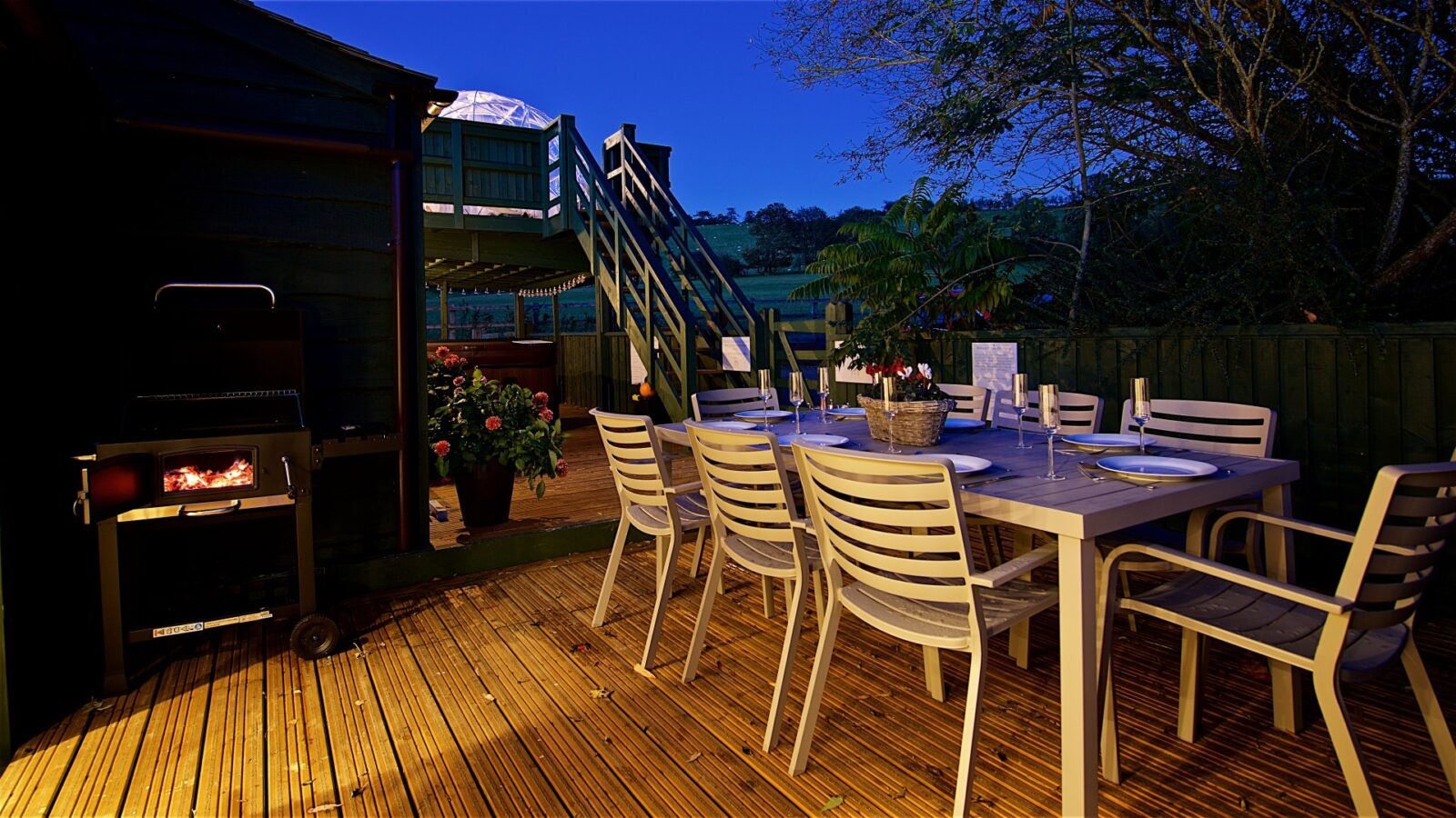 The Georges Getaway outdoor dining setup features a charming wooden deck at dusk, complete with a table, chairs, and a lit grill nearby, all surrounded by lush trees and plants.